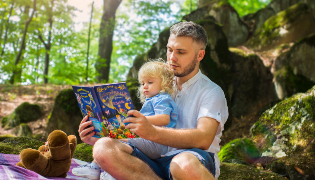 A man reads a colorful book to a young child on a blanket in Wisconsin woods, symbolizing family financial planning outdoors.