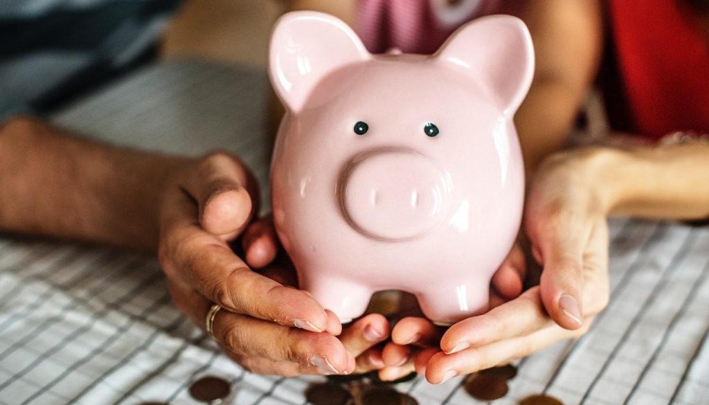 Two pairs of hands hold a pink piggy bank over scattered coins, symbolizing Wisconsin tax planning with fiduciary financial advisors.