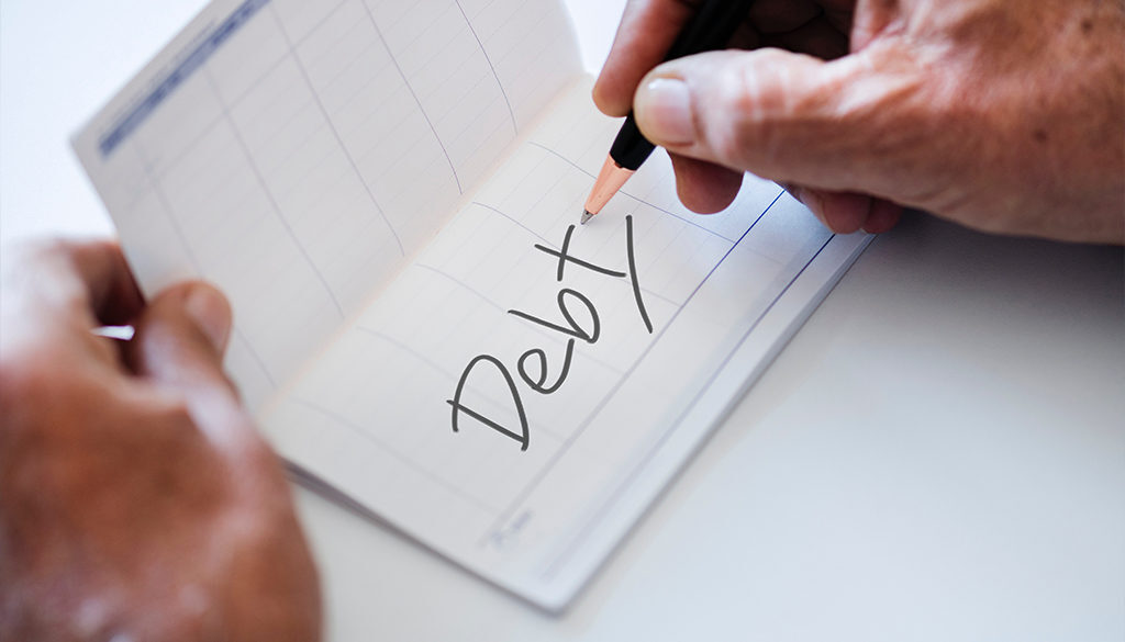A person writes "Debt" in a checkbook register with a pencil, tracking accounting services from fiduciary advisors in Wisconsin.