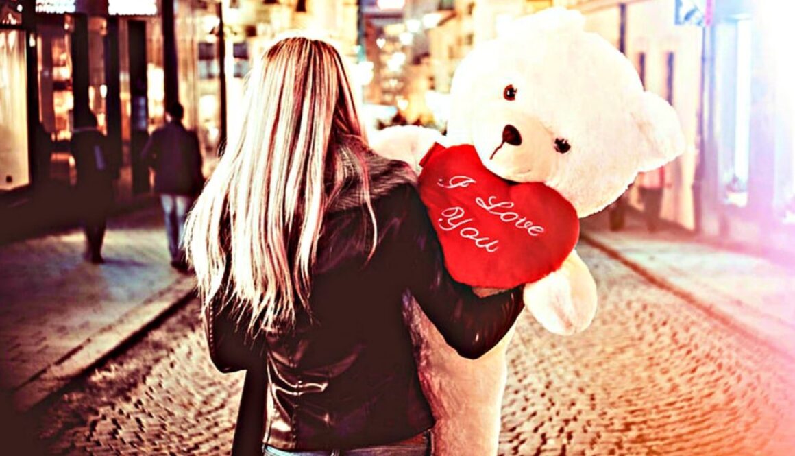 A woman with long hair holds a white teddy bear with a red heart on a cobblestone street in Wisconsin.