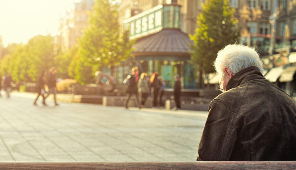 An older man with white hair sits alone on a bench in a sunlit Wisconsin city square, symbolizing thoughtful financial planning.