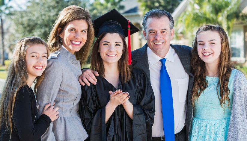 A family consisting of a mother, father, and three daughters smile. One of the daughters is wearing a graduation cap and gown.