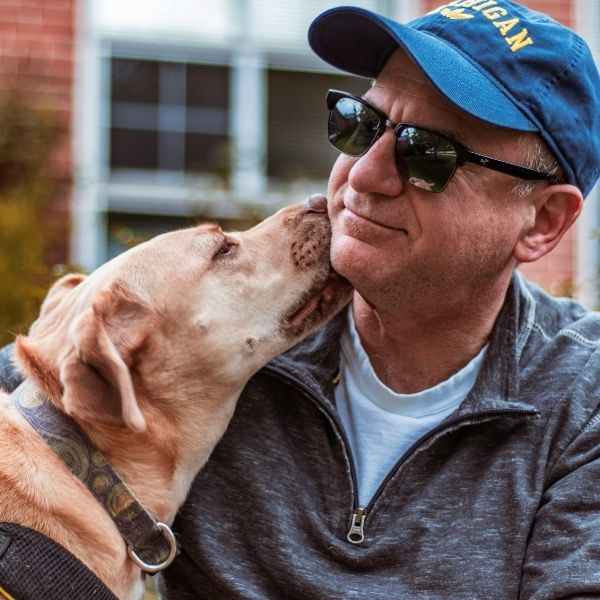 A man in sunglasses and a blue cap smiles as his yellow dog licks his chin outdoors in Wisconsin, reflecting local community values.