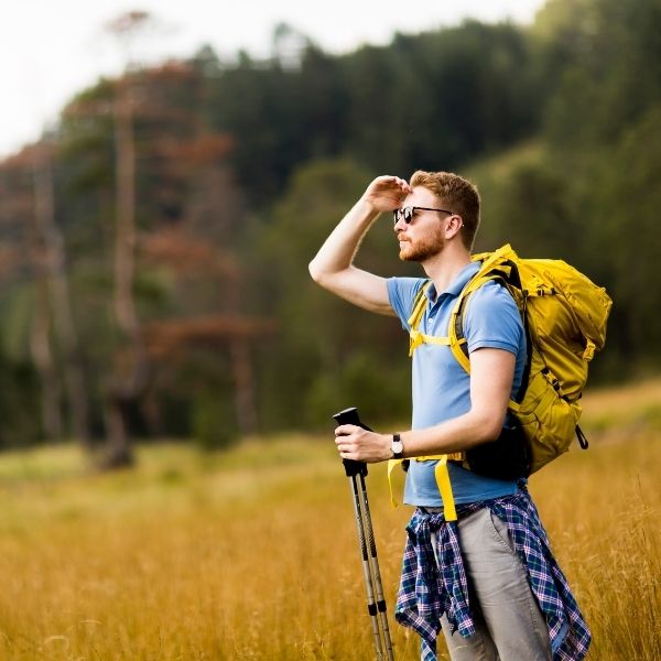 A man with a yellow backpack and hiking poles stands in a grassy Wisconsin field, representing financial planning outdoors.