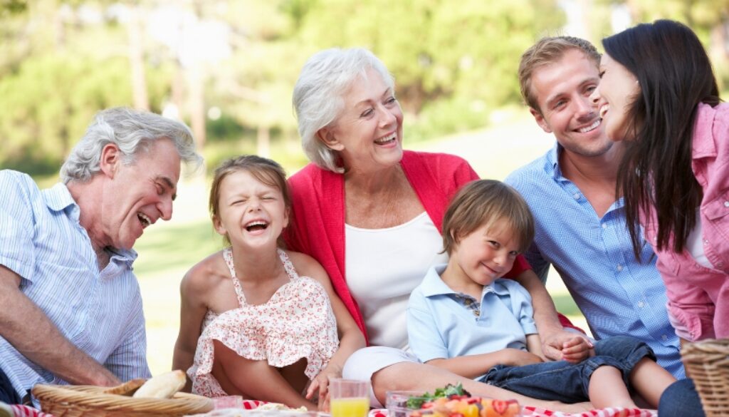 Grandparents, parents, and young children smiling and laughing while enjoying a summer picnic.
