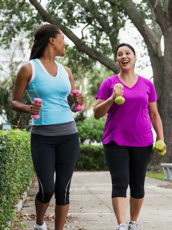 Two women walk on a scenic Wisconsin path, smiling with small hand weights, enjoying wellness—like Advisors Management Group clients.
