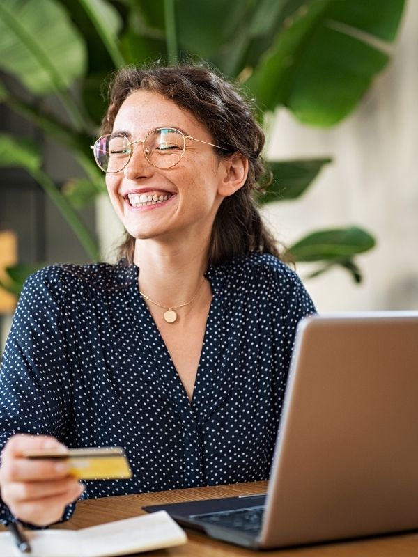 A smiling woman with glasses holds a credit card at a desk with a laptop and notepad, highlighting financial planning in Wisconsin.