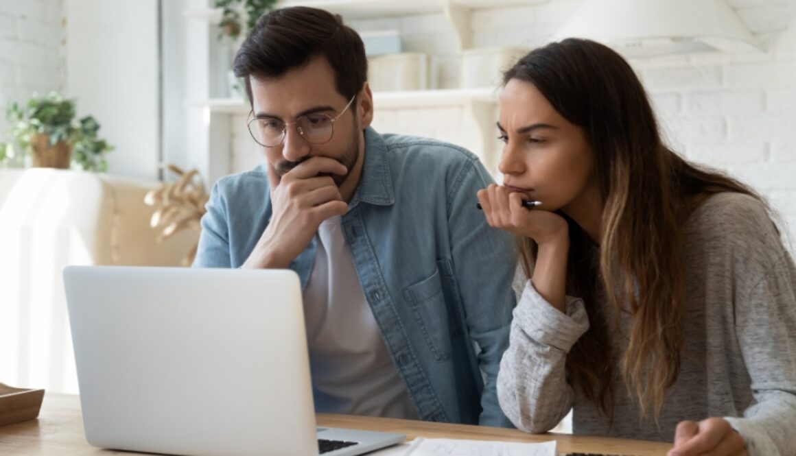 A young couple sit contemplatively on the couch looking at a laptop.