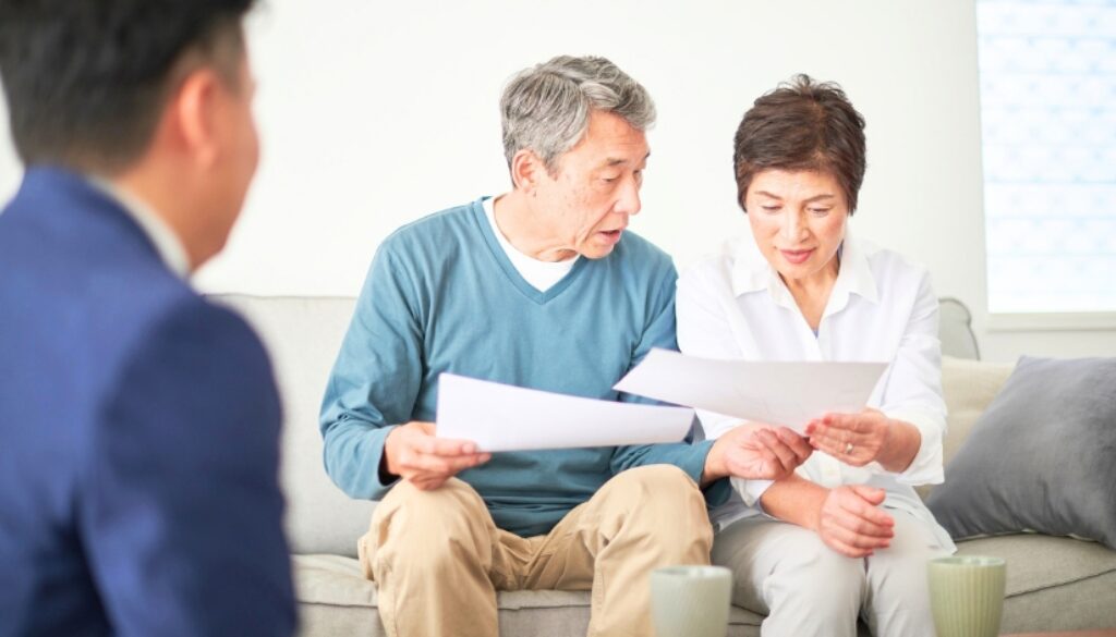 An elderly couple sitting on their couch reviewing paperwork. A man in a suit is blurred to the side.