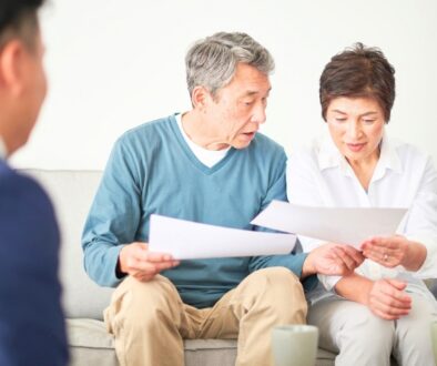 An elderly couple sitting on their couch reviewing paperwork. A man in a suit is blurred to the side.