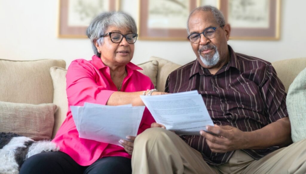 An elderly couple sitting on their couch reviewing and pointing to paper documents.