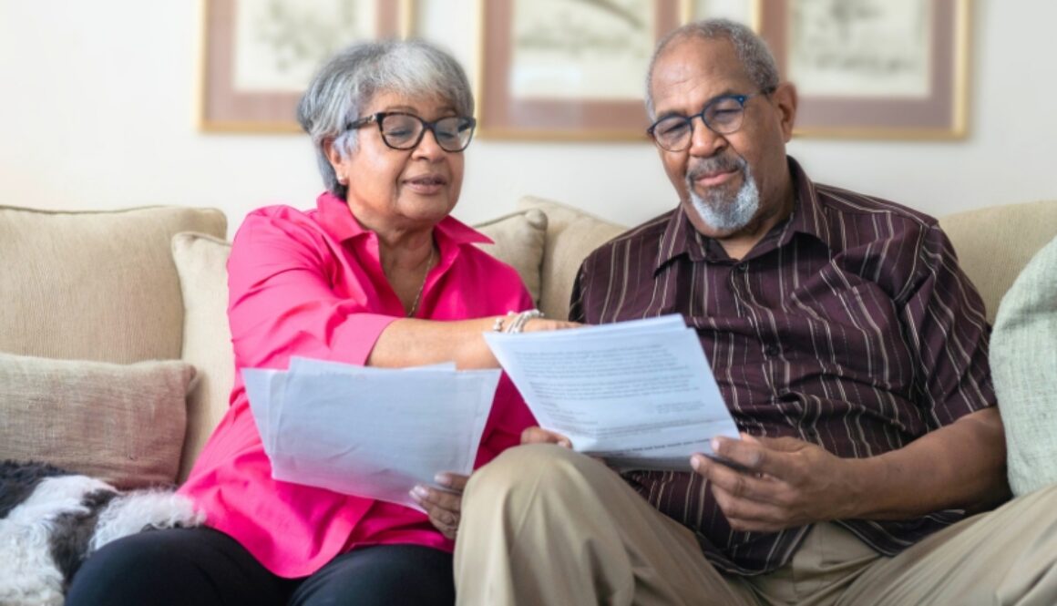 An elderly couple sitting on their couch reviewing and pointing to paper documents.