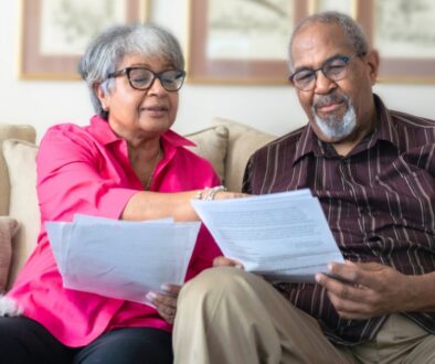 An elderly couple sitting on their couch reviewing and pointing to paper documents.