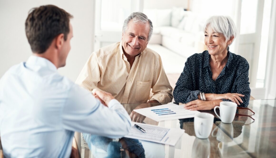 An older couple sitting at a dining table shaking hands with a professional. Paperwork and coffee cups sit on the table.