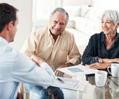 An older couple sitting at a dining table shaking hands with a professional. Paperwork and coffee cups sit on the table.