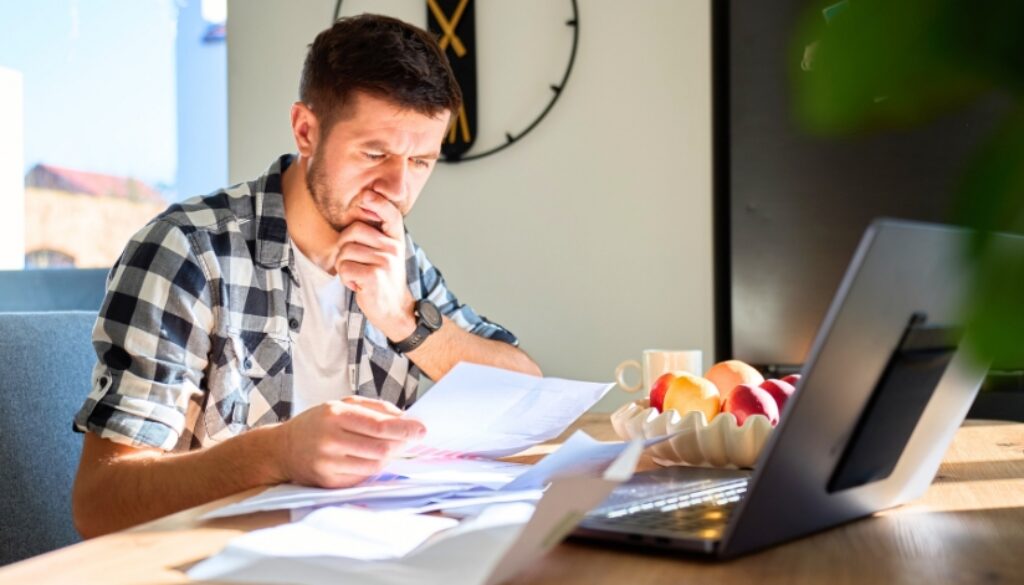 A young professional reviewing paperwork at his kitchen table. A laptop sits open in front of him.