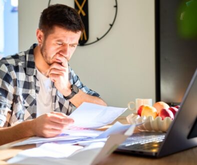 A young professional reviewing paperwork at his kitchen table. A laptop sits open in front of him.