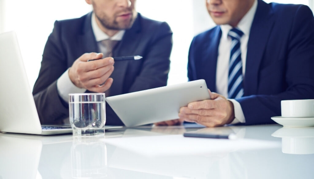 Men in suits sitting in a conference room looking at a tablet.