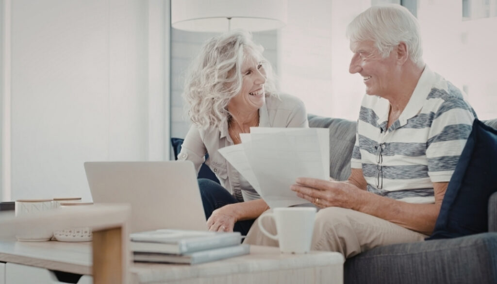 Older couple smiling while reviewing paperwork together on a couch with a laptop on the table.