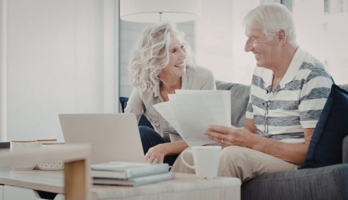Older couple smiling while reviewing paperwork together on a couch with a laptop on the table.
