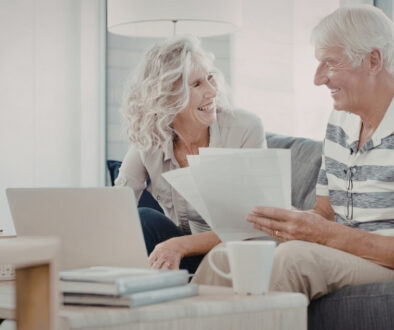 Older couple smiling while reviewing paperwork together on a couch with a laptop on the table.