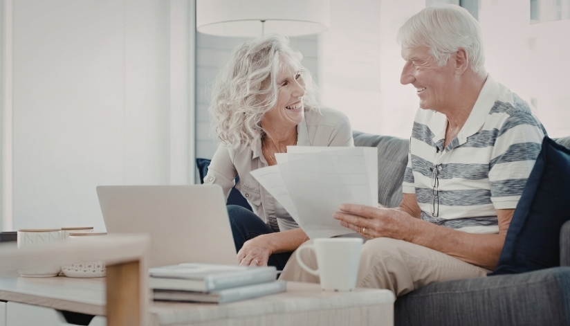 Older couple smiling while reviewing paperwork together on a couch with a laptop on the table.